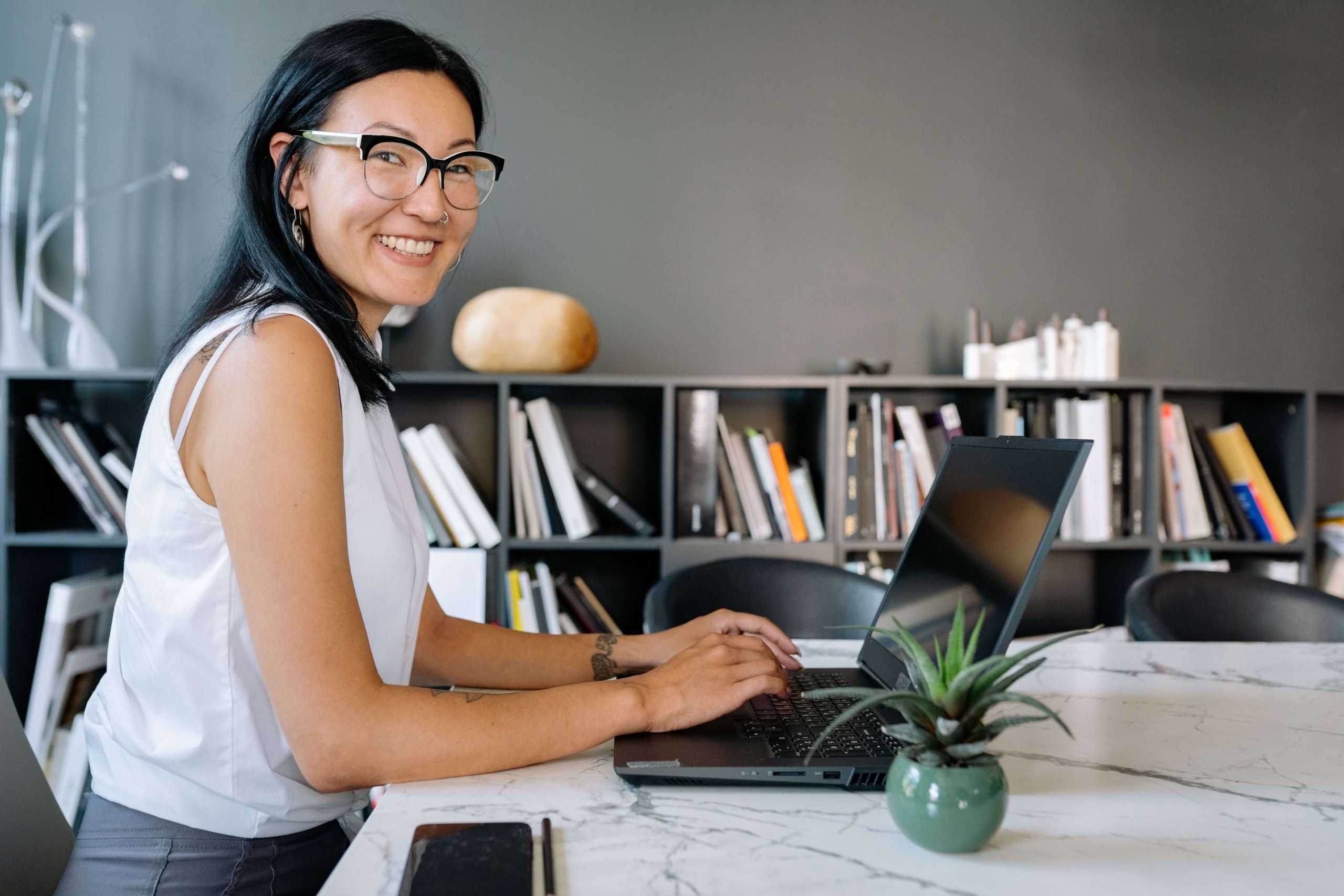 smiling woman working on laptop | keywords for online counseling pages | seo services for therapists | seo company for counselors | Canada | Maine | Vermont | Massachusetts A smiling woman with dark hair, glasses, and a nose ring sits at a marble desk with a laptop and small plant, turning toward the camera in front of a dark bookshelf. Your voice is what makes clients choose you, and the right SEO for therapy practice strategy makes sure that voice is what shows up online.