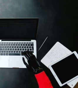 A robotic hand types on a laptop keyboard next to a notepad and tablet. This image represents the risk of relying solely on AI to generate content — without human oversight, private practice SEO content can lack the accuracy, voice, and nuance that potential clients need to feel confident reaching out. 