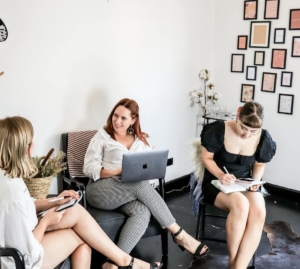 Three women collaborating in a bright office, one on a laptop and others taking notes, representing how following SEO best practices and reviewing content together helps teams catch AI hallucinations before publishing.