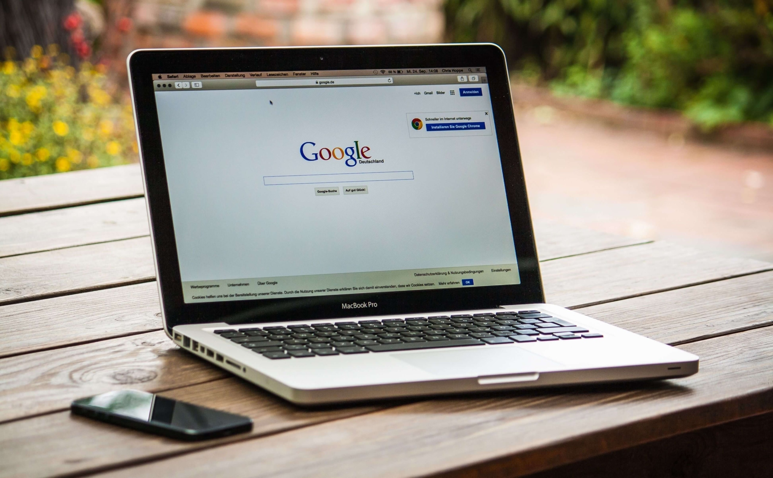 Google homepage open | EEAT for therapists | content credibility for therapy websites | expertise authority trustworthiness | Arkansas | Montana | Kansas A MacBook Pro laptop sits open on a wooden outdoor table displaying the Google homepage with a smartphone placed beside it and greenery visible in the background. Understanding what Google looks for when it evaluates your therapy website is the first step toward building an online presence that actually works for your practice.