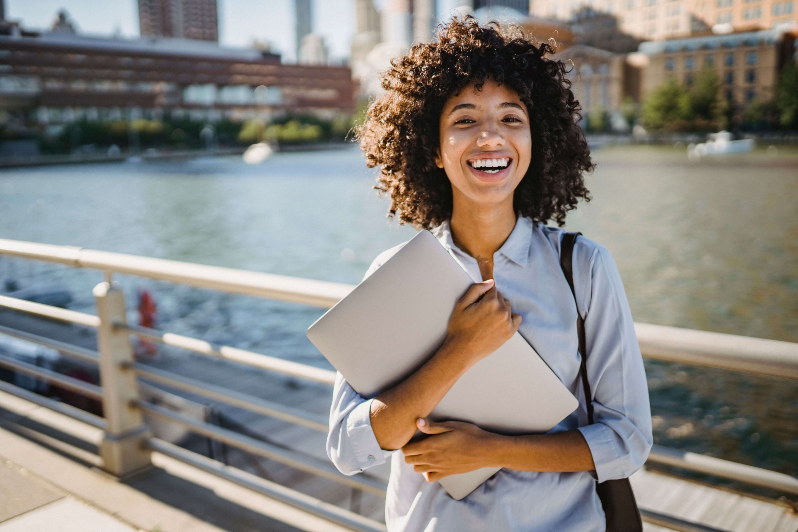 smiling woman holding laptop | EEAT for mental health professionals | seo for private practices | expertise authority trustworthiness | Massachusetts | Maine | Vermont | Arkansas A smiling woman with curly hair holds a laptop against her chest while standing outdoors on a bridge with a river and city buildings visible behind her. When your therapy website reflects your real expertise and experience, it builds the kind of trust that turns a casual visitor into a client who actually reaches out.