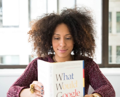 Woman reading a book titled 'What Would Google Do?' at a desk, representing how understanding Google's AI content policy and AI content SEO helps you create content that ranks well.